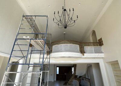 A large, high-ceilinged room under renovation with scaffolding, an ornate chandelier, covered railings, and plastic sheeting protecting the staircase and floors.